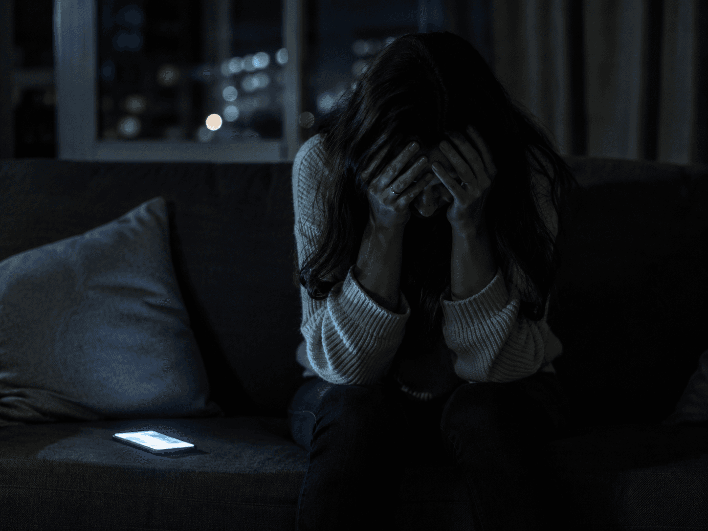 A lonely woman sitting in a dim room beside a glowing phone.